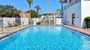 A rectangular swimming pool with a clear blue water surface, surrounded by a white fence, and adjacent to a building with a balcony.