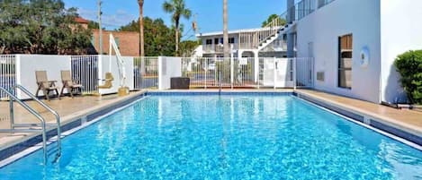 Une piscine rectangulaire à l’eau d’un bleu limpide, entourée d’une clôture blanche et jouxtant un bâtiment doté d’un balcon.