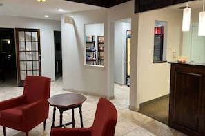 Um saguão de hotel com um balcão de recepção, uma mesa redonda pequena e duas poltronas vermelhas.