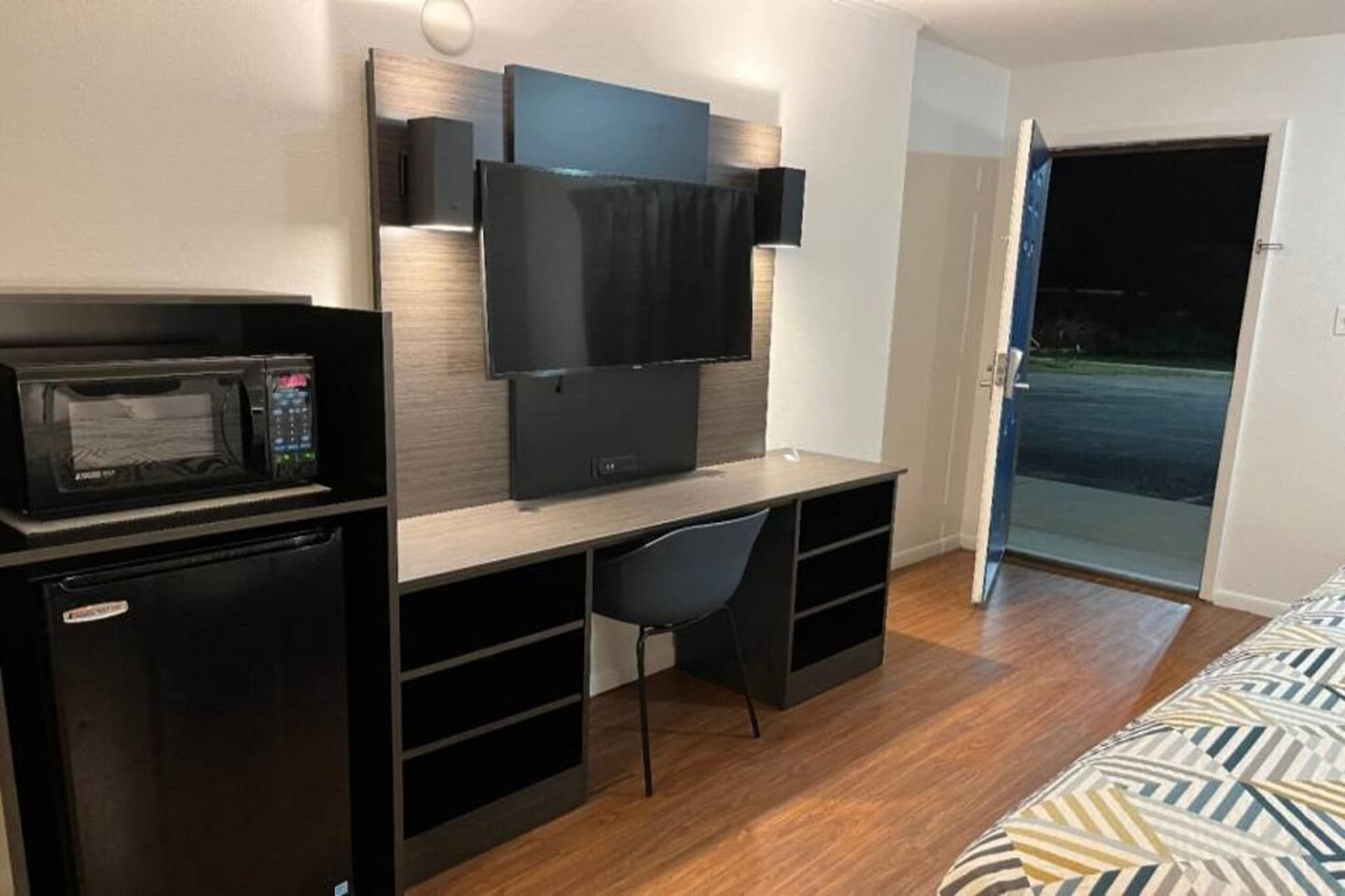 A compact room with a kitchenette, a flat-screen TV mounted on the wall, a single chair, and a bed with a patterned cover.