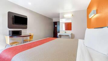 Habitación de hotel con cama, escritorio, silla, televisor y baño con espejo y lavamanos.