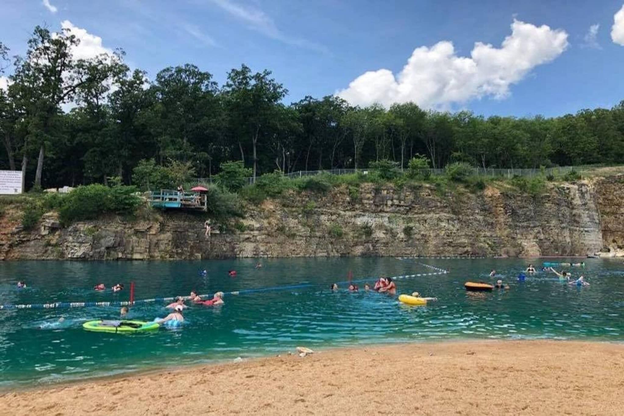 A beach with clear water, people swimming and floating, and a rocky cliff with trees.