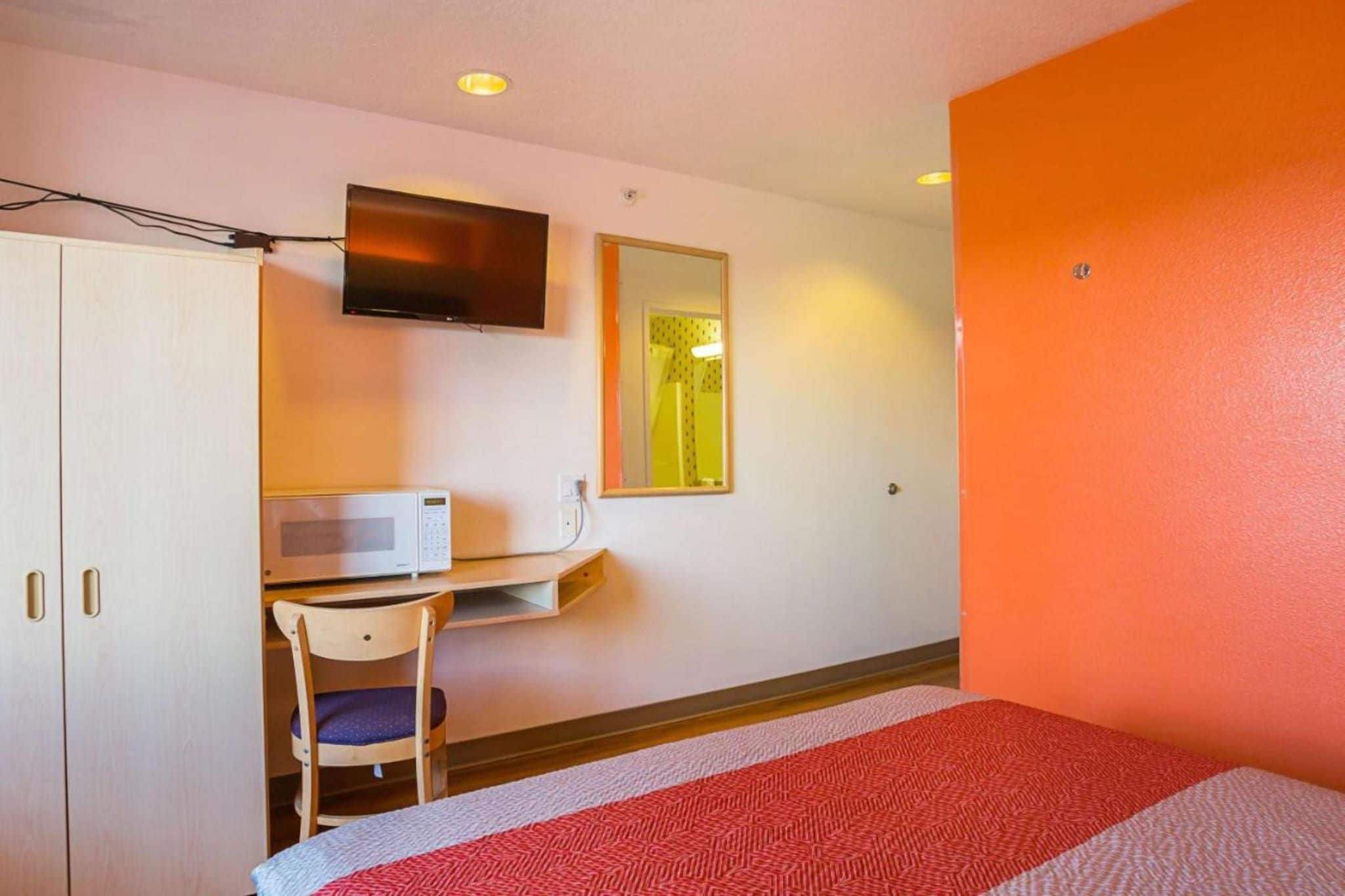 A hotel room with a bed, a desk with a chair, a microwave, a television, and a mirror.