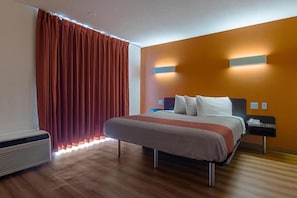 Ein Hotelzimmer mit Bett, Nachttisch, Lampe, Heizung und einem Fenster mit Vorhängen.