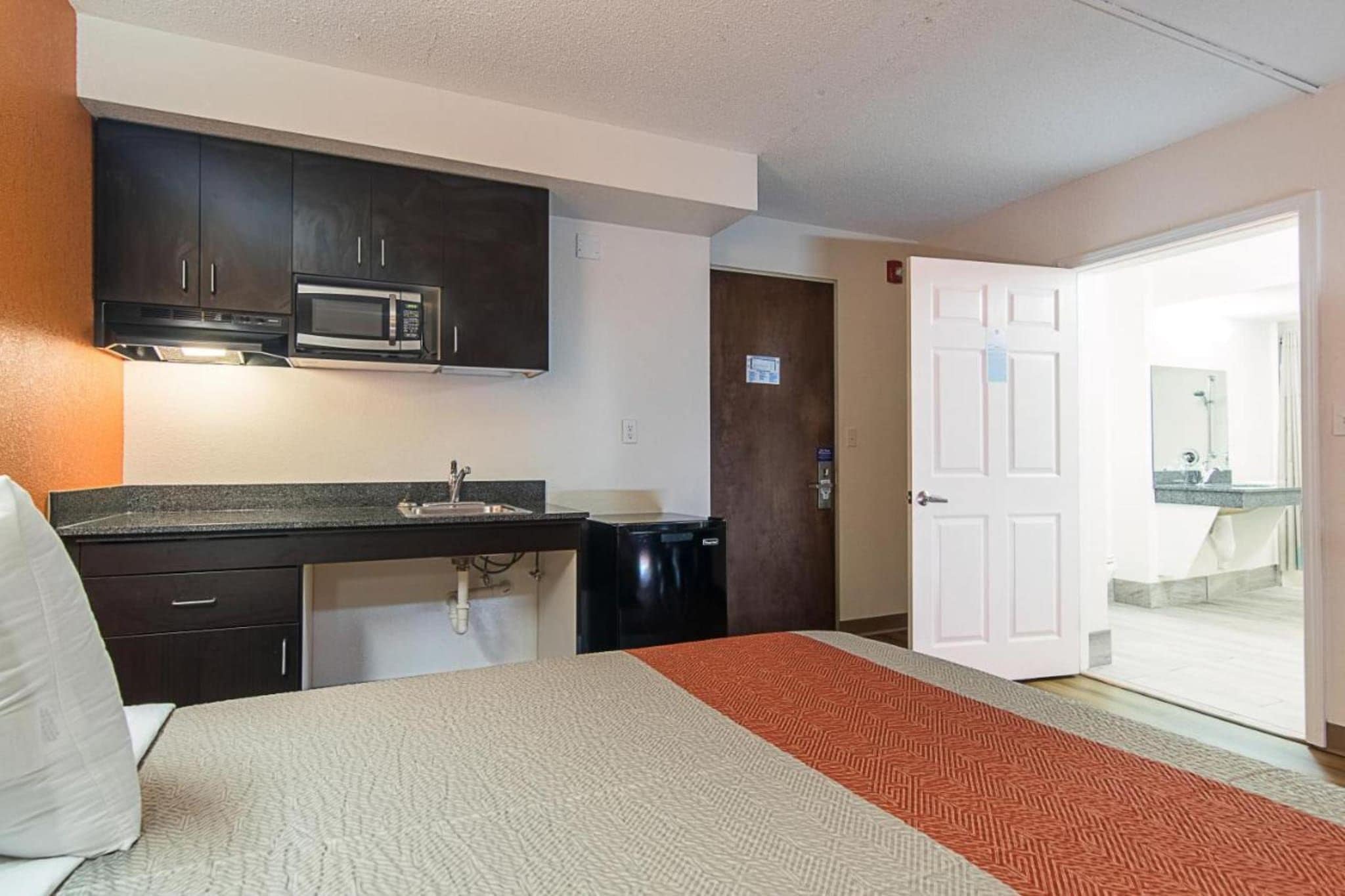 Une chambre d’hôtel compacte comprenant un coin cuisine, un lit et une salle de bain visible à travers une porte ouverte.