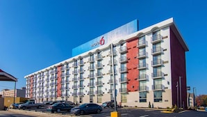 Un hotel de varios pisos con un llamativo letrero de 'Motel 6', varios balcones y un estacionamiento con varios automóviles.