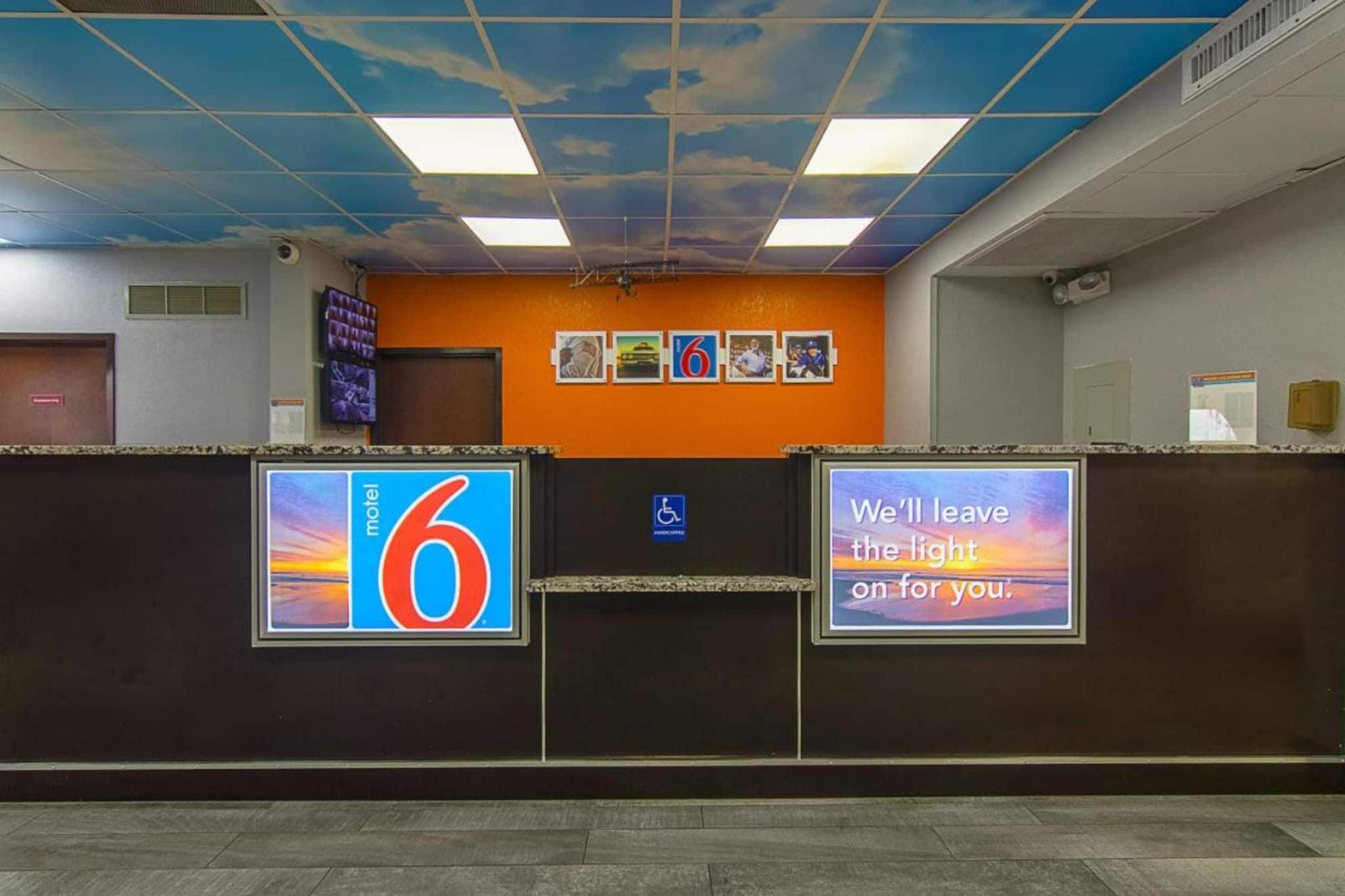 Un espace d’accueil d’hôtel avec un plafond bleu, un panneau indiquant le numéro 6, un symbole indiquant l’accessibilité aux personnes à mobilité réduite et deux écrans numériques affichant des messages.