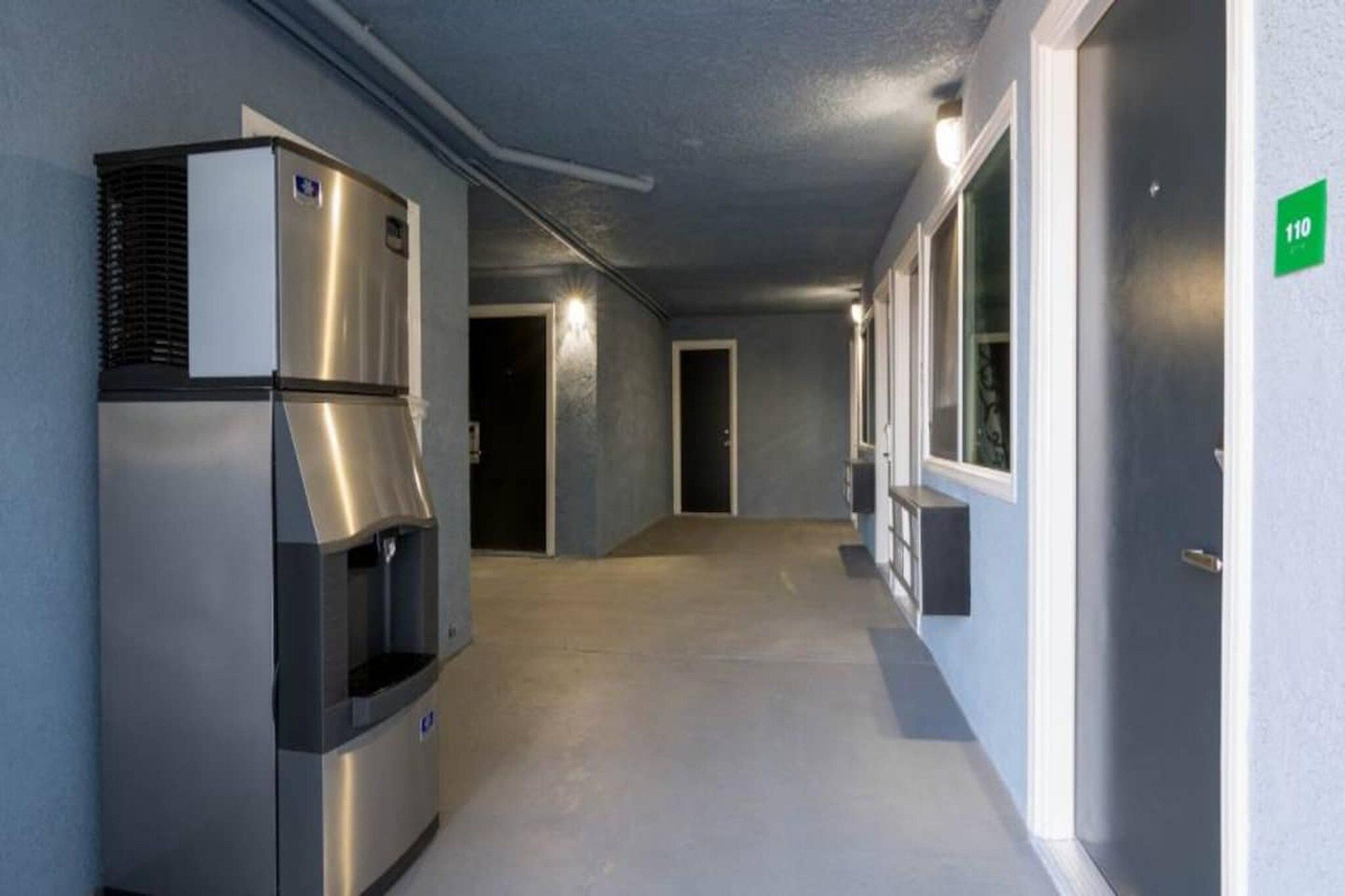 A corridor with a stainless steel refrigerator and a water dispenser, a door with a green sign, and a window on the right wall.