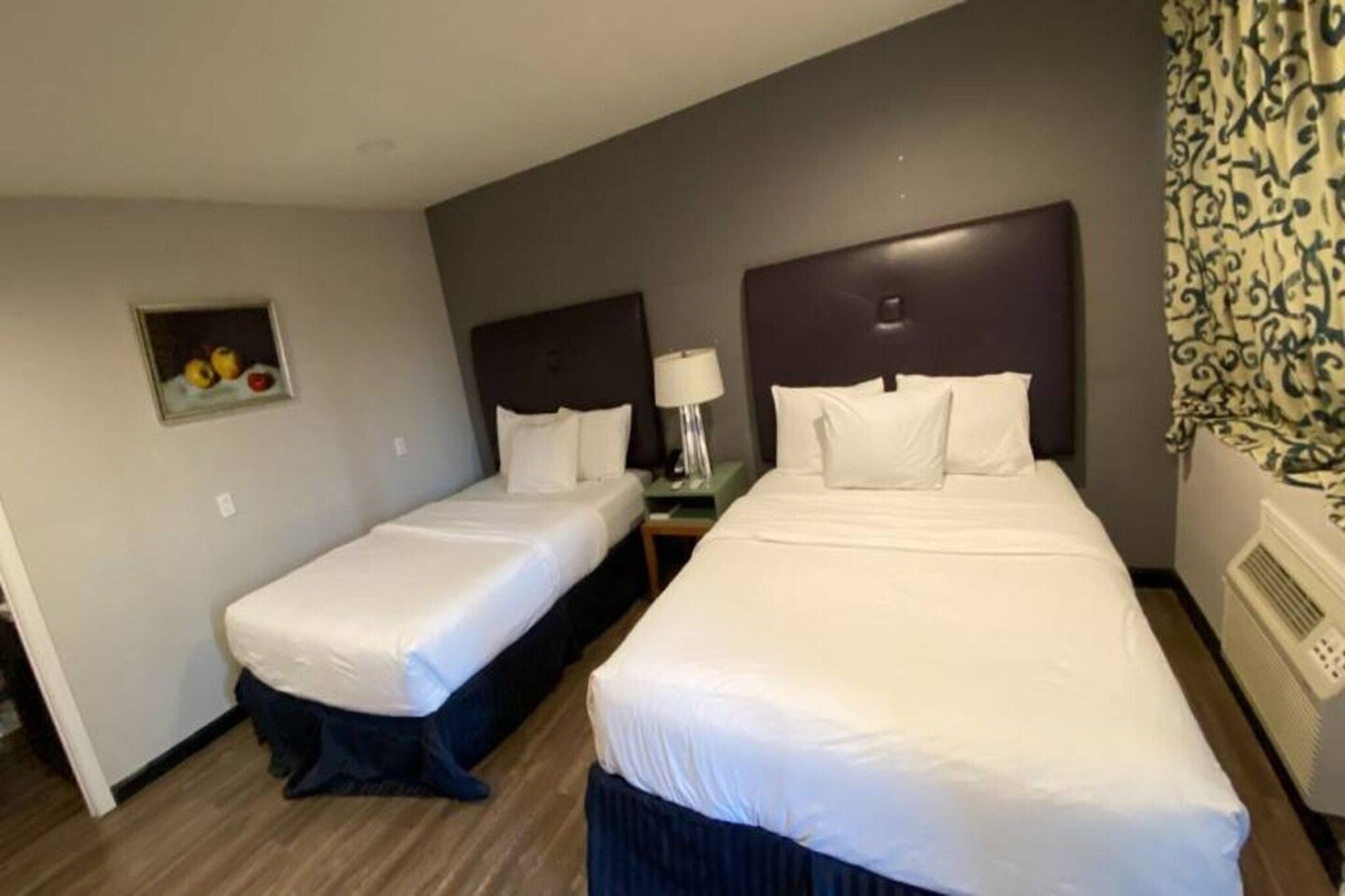 A hotel room with two beds, a nightstand, a lamp, and a wall-mounted air conditioner.