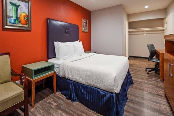 A hotel room with a bed, a desk, a chair, and a painting on the wall. - Spacious 2-Bed Queen|Fridge|Pool (Ridgecrest CA)