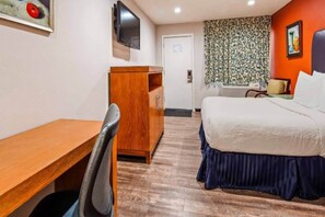 A hotel room with a wooden desk, a bed with a blue skirt, a television on the wall, and a patterned curtain.