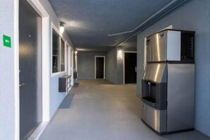 A corridor with a stainless steel refrigerator, a door with a green sign, and a window on the left.