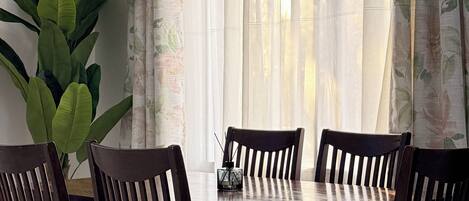 A dining area with a table and chairs, a potted plant, and a window with sheer curtains.