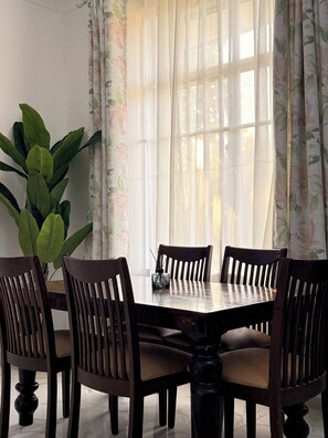 Un coin repas avec une table et des chaises, une plante en pot et une fenêtre ornée de rideaux légers.