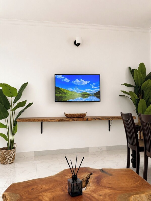 Un salon moderne avec un téléviseur à écran plat fixé à un mur blanc, une table basse en bois et deux plantes en pot.