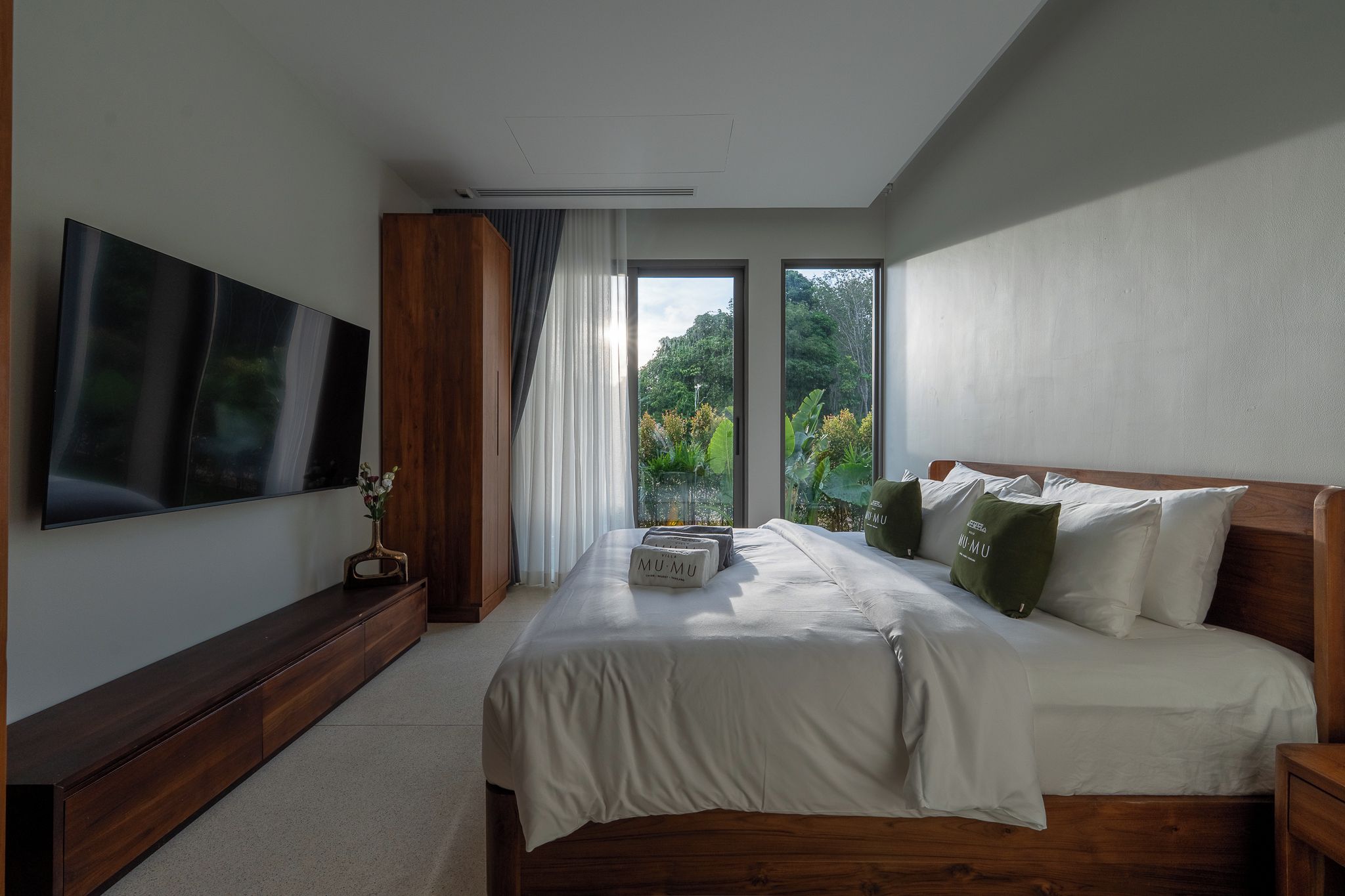 A modern hotel bedroom with a large bed, a flat-screen TV, and a view of greenery through the window.
