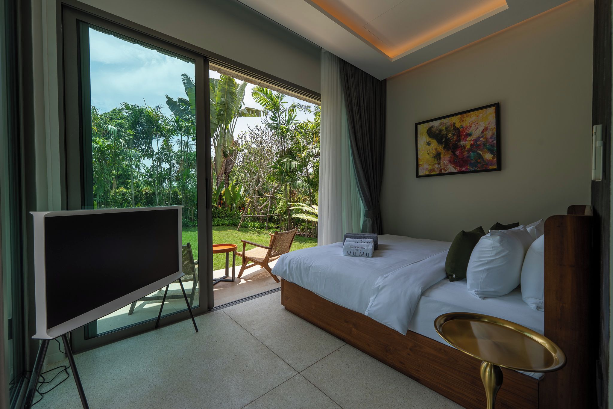 A hotel room with a large bed, a TV, a dining area with chairs, and a view of greenery outside.