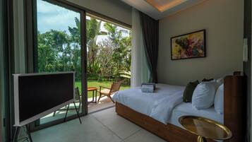 A hotel room with a large bed, a TV, a dining area with chairs, and a view of greenery outside.