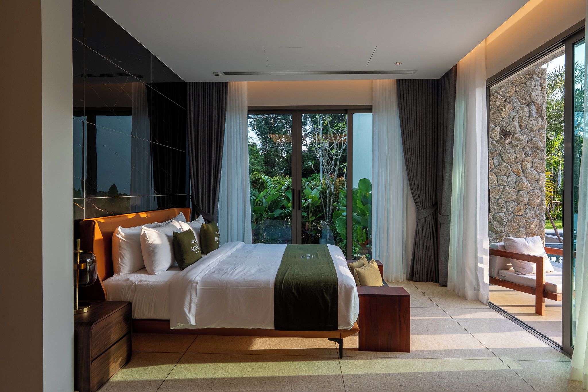 A modern bedroom with a large bed, a nightstand, a wooden bench, and a view of greenery through large windows.