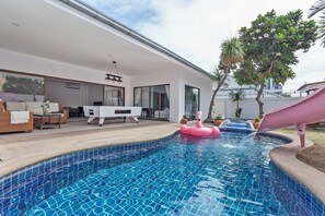 Ein Schwimmbecken mit einem rosa Luftmatratze, ein weißer Außenbereich zum Sitzen und ein modernes Haus mit großen Fenstern.