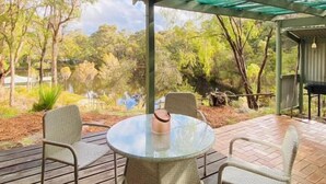Un patio exterior con una mesa redonda y sillas, rodeado por una terraza de madera y una zona cubierta con vistas a árboles y una masa de agua.