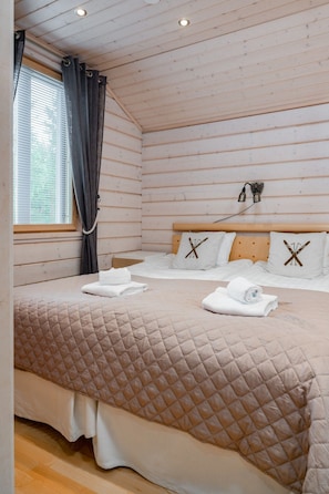 A bedroom with a wooden ceiling, a bed with a quilted cover, two pillows with a ski design, and a window with curtains. - Villa Hutiputti A · Pool · Ski & Golf (Kittilä)