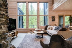 A cozy living room with a stone wall, large windows, a flat-screen TV, a sofa, a coffee table, and a fireplace. - Villa Hutiputti A · Pool · Ski & Golf (Kittilä)