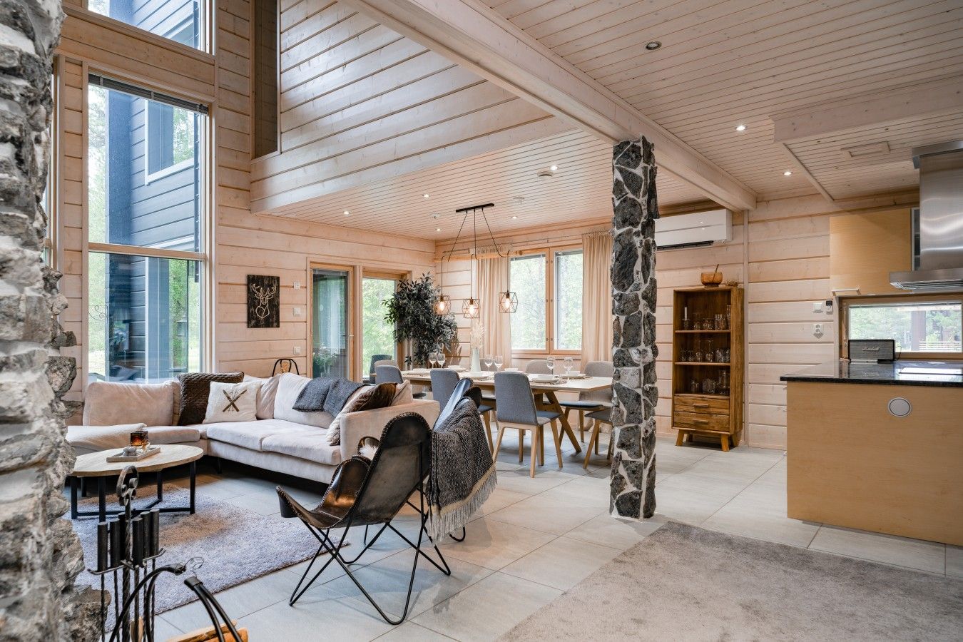 Een ruime woonkamer met een bank, stoelen, een eettafel en een keukenhoek met een stenen kolom en houten keukenkastjes.