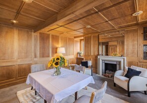 Ein Zimmer mit holzgetäfelter Wand, einem Kamin, einem Esstisch mit weißem Tischlein und einer Couch mit Kissen.