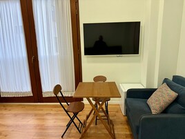 A room with a wooden table and chairs, a sofa, a TV mounted on the wall, and a sliding door with curtains.