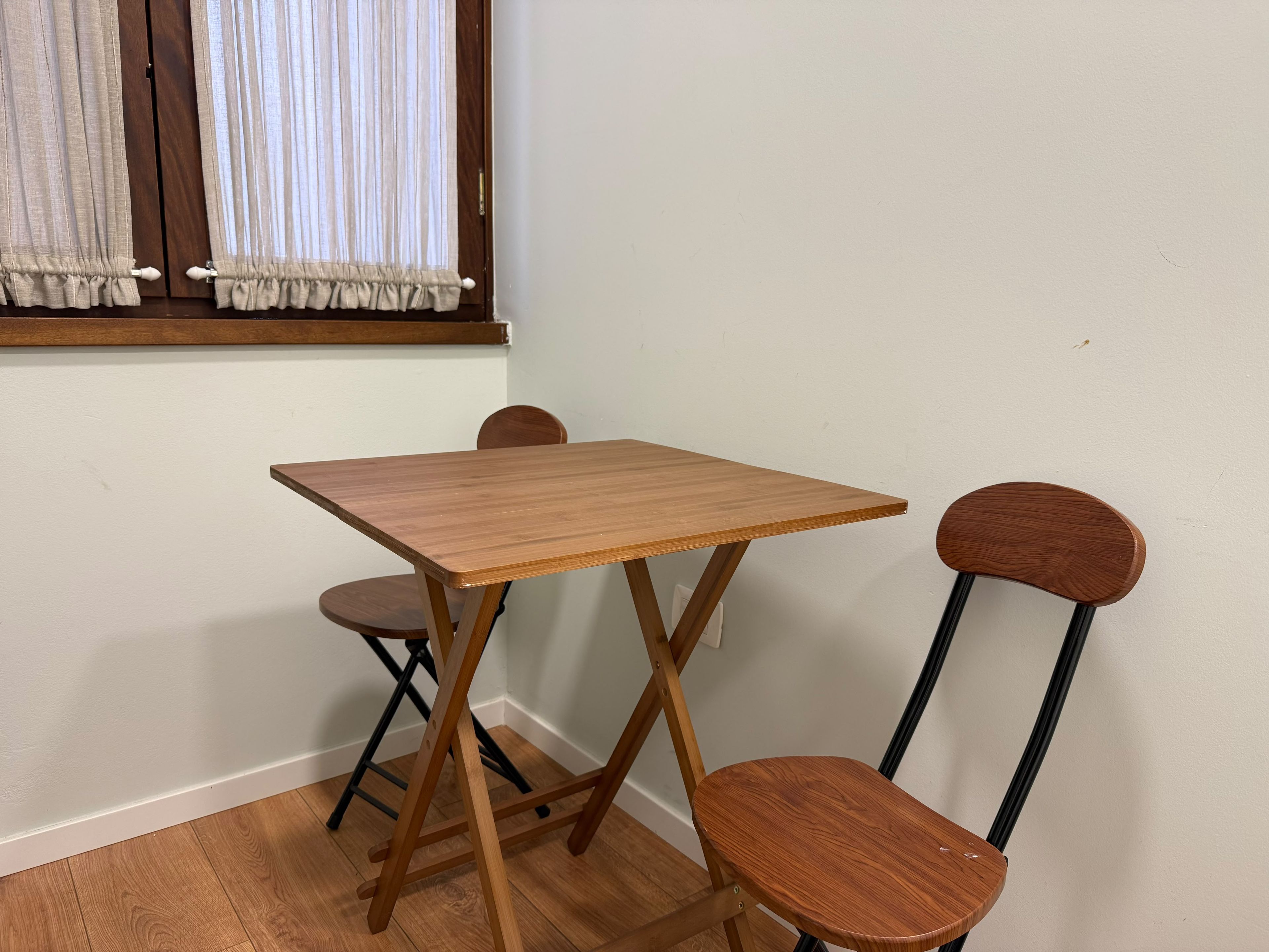 Un petit coin repas avec une table en bois et deux chaises, une fenêtre avec des rideaux et un mur sobre.