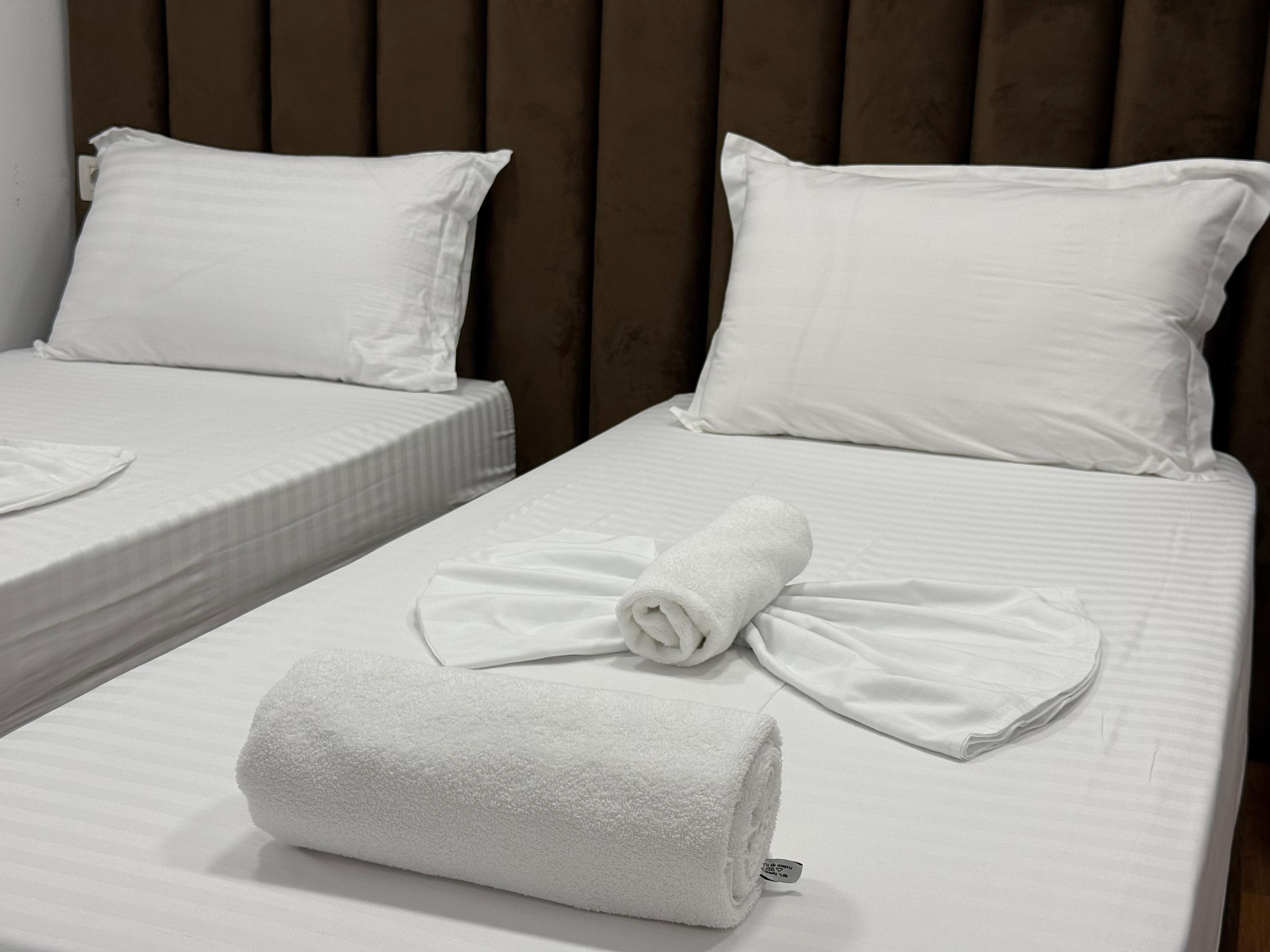 Two single beds with white linens and pillows, a rolled white towel on the bed, and a wooden headboard.