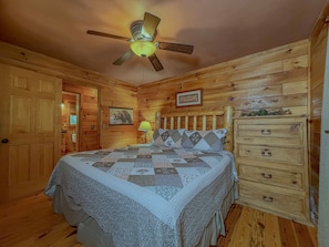 A wooden cabin bedroom with a bed, a nightstand, a dresser, a ceiling fan, and a framed picture on the wall. - Cozy 1BR Loft Cabin w/hot tub, fire pit | Sleeps 4 (Sautee Nacoochee)