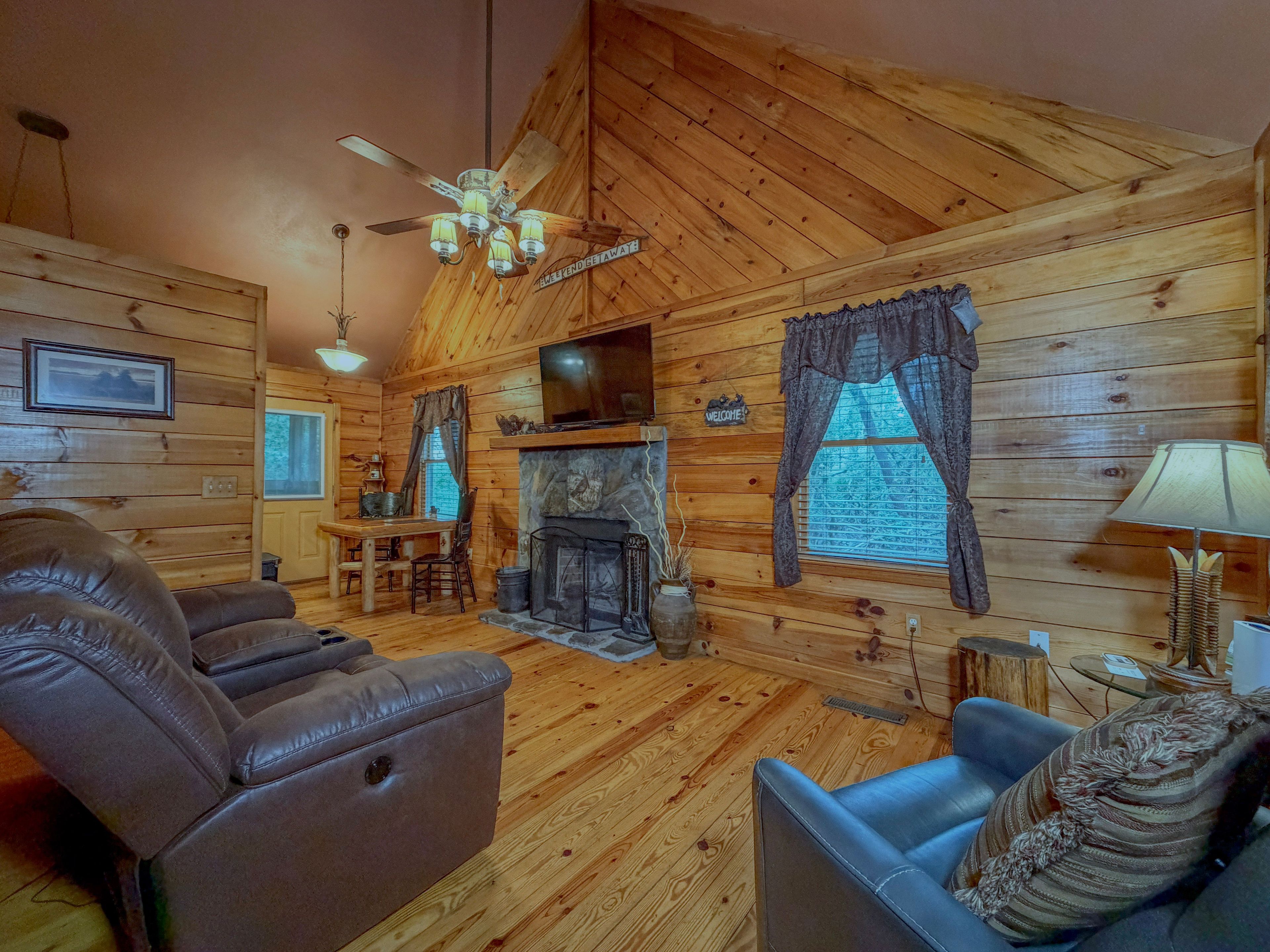 A log cabin interior with a fireplace, leather sofas, a dining table, and a television.