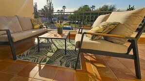 Ein sonniger Außenbereich mit Sitzgelegenheiten, darunter eine Couch, ein Sessel und ein Couchtisch, mit einem gemusterten Teppich und Blick auf die Umgebung.