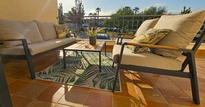Ein sonniger Außenbereich mit Sitzgelegenheiten, darunter eine Couch, ein Sessel und ein Couchtisch, mit einem gemusterten Teppich und Blick auf die Umgebung.