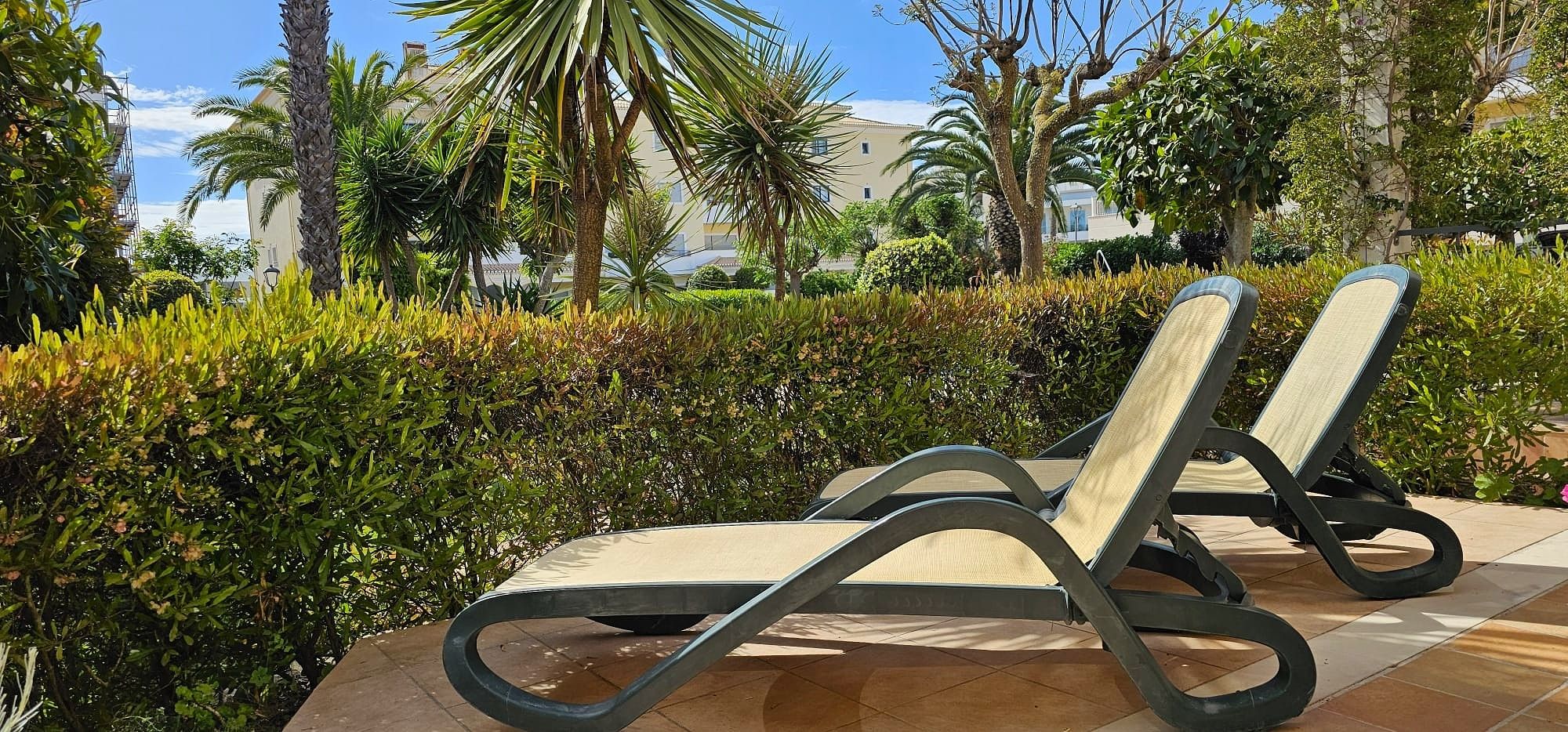 Two lounge chairs with a cushioned seat and backrest, positioned on a tiled patio area with a backdrop of well-maintained hedges and palm trees.