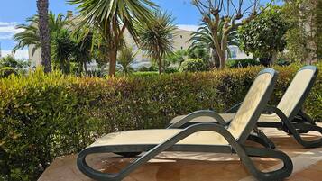 Two lounge chairs with a cushioned seat and backrest, positioned on a tiled patio area with a backdrop of well-maintained hedges and palm trees.