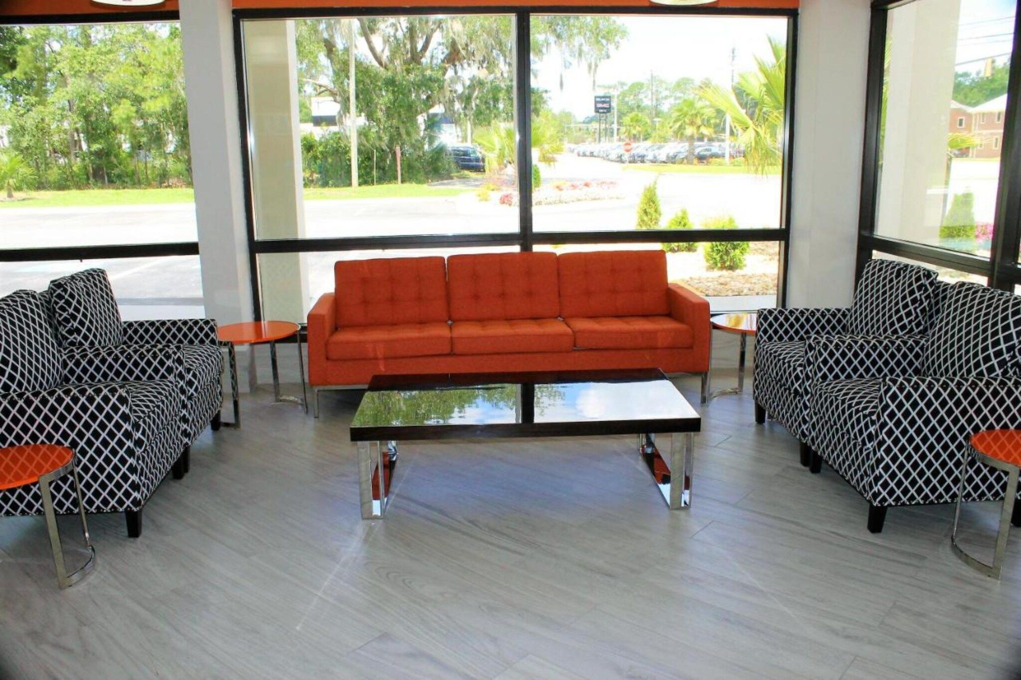A seating area with a sofa and patterned chairs, a coffee table, and large windows with a view of a parking lot and greenery.