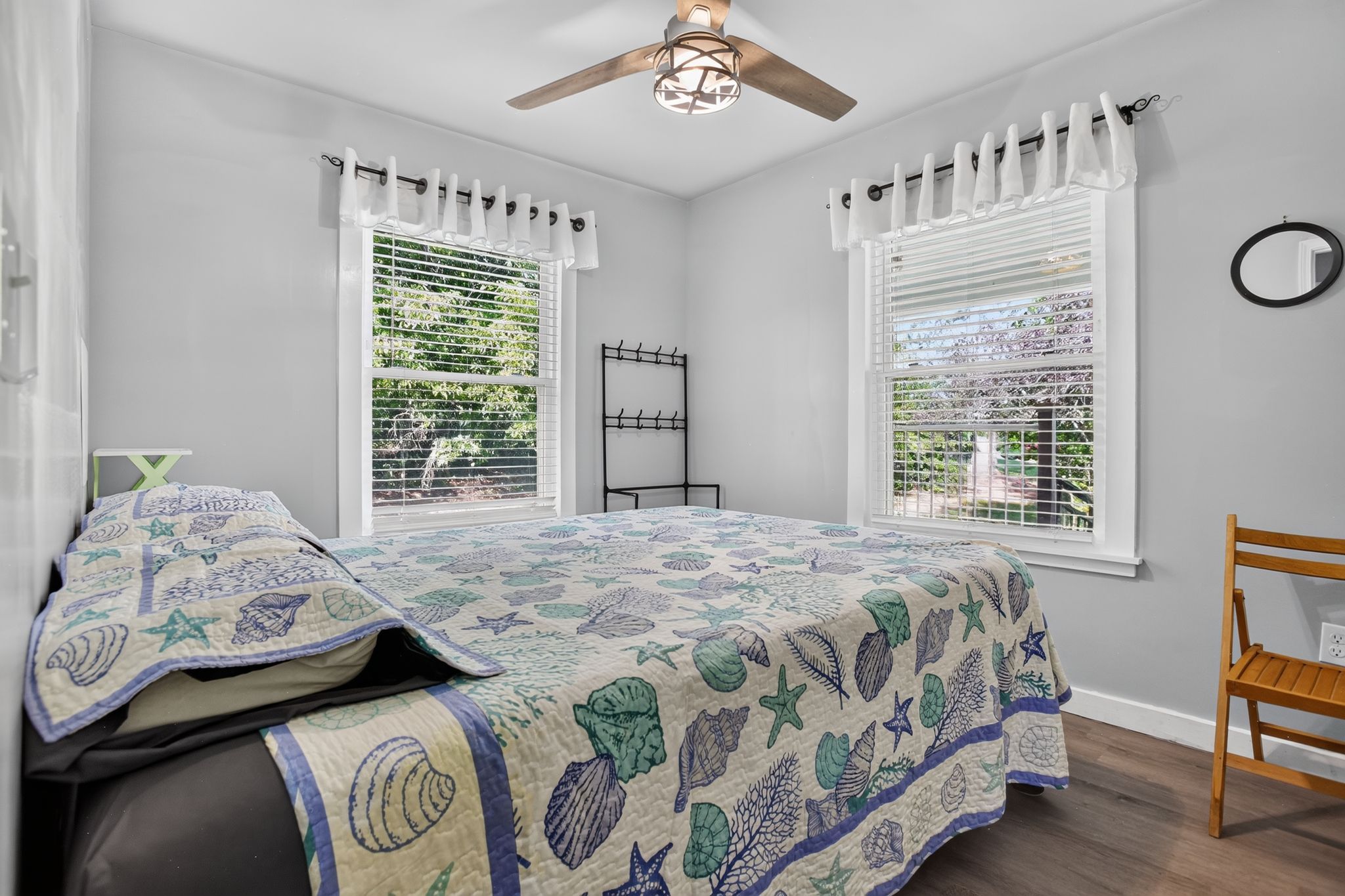 A bedroom with a bed, a wooden chair, a ceiling fan, and two windows with curtains.