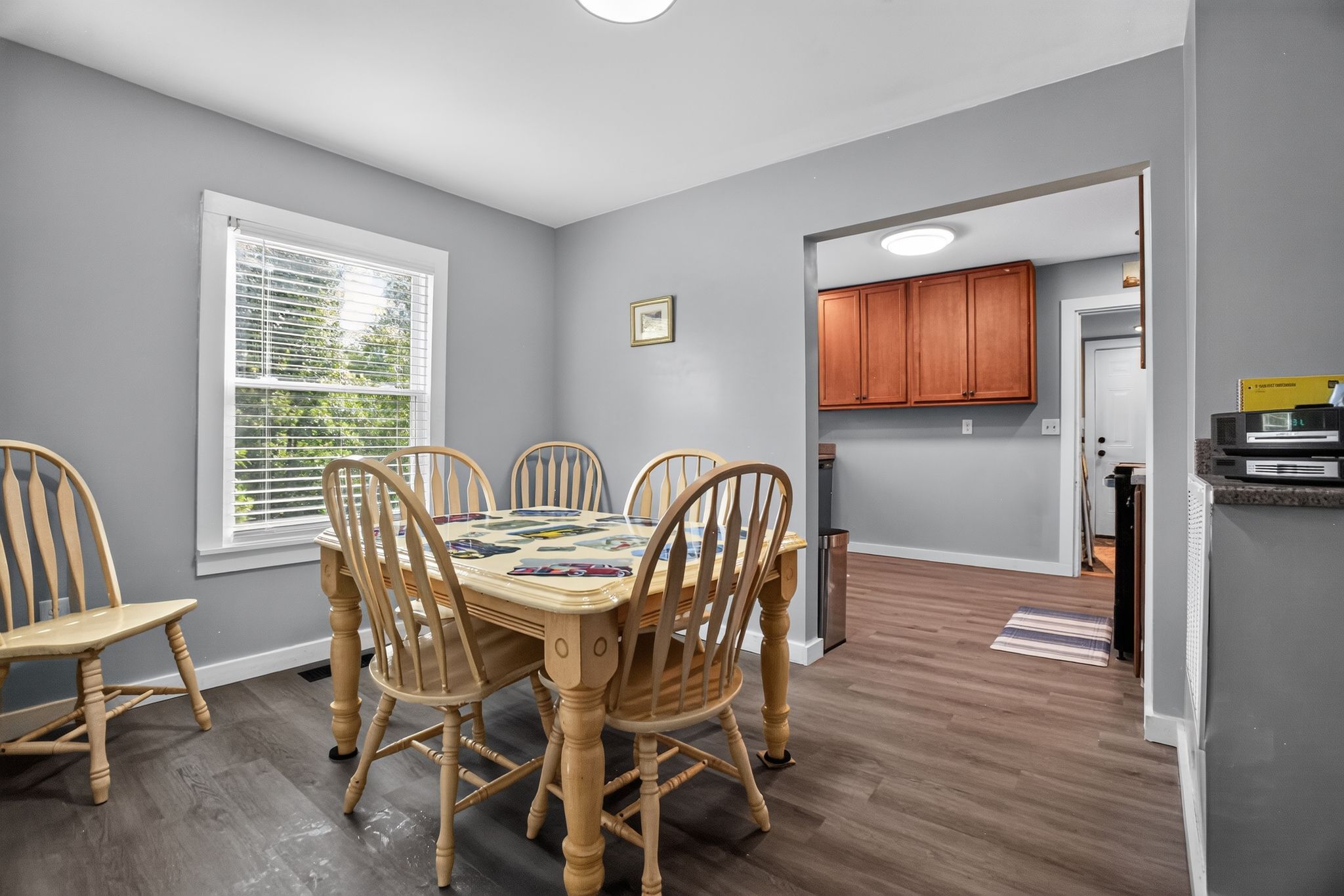 A dining area with a wooden table and chairs, a window with blinds, and a kitchen area with wooden cabinets.
