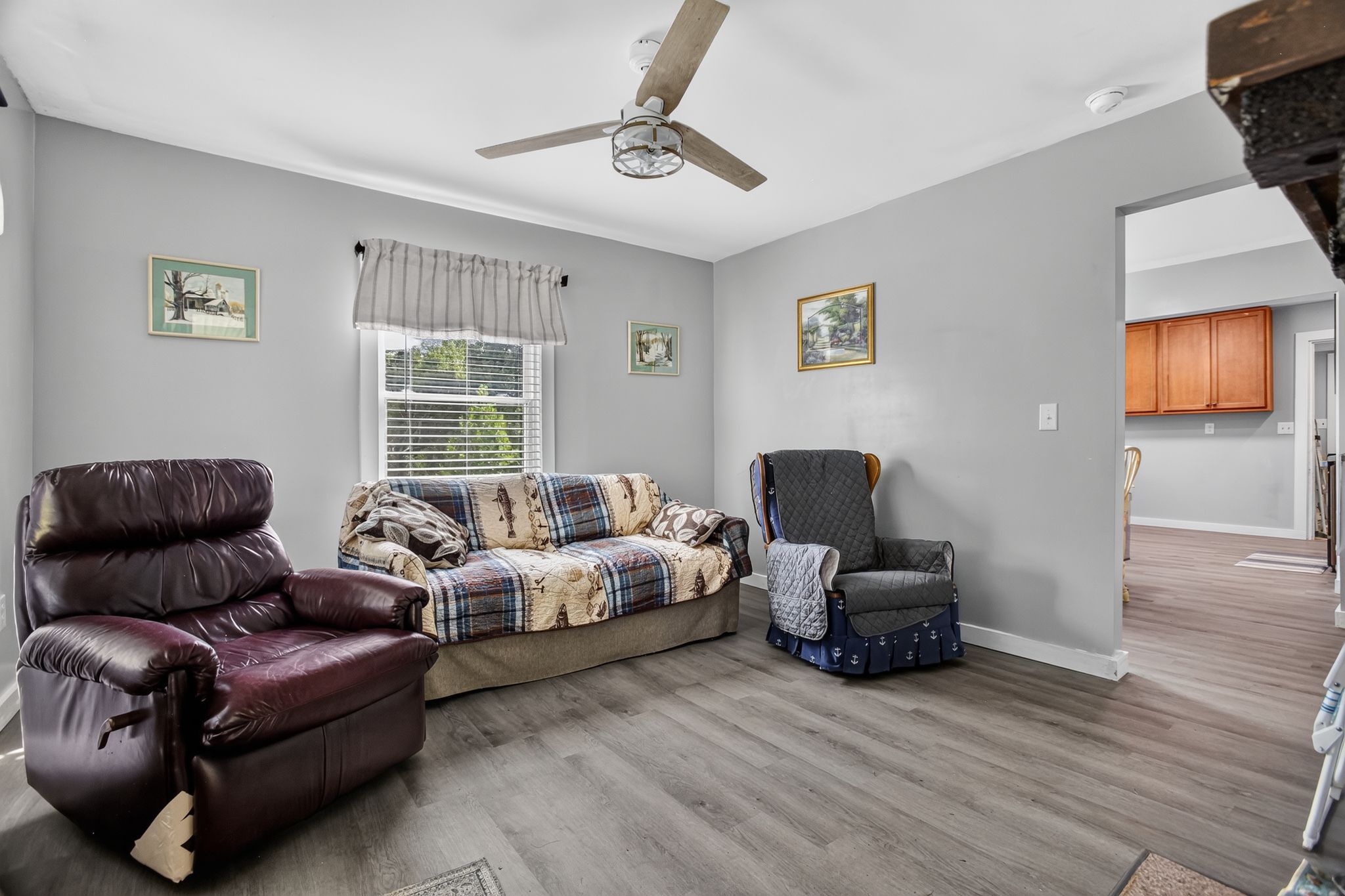 A living room with a ceiling fan, a leather armchair, a sofa with a patterned blanket, a quilted chair, and a window with blinds.