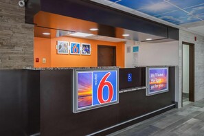 Een hotel receptie met een opvallend bord met het getal '6', een symbool voor rolstoeltoegankelijkheid en een digitaal scherm waarop een bericht staat.