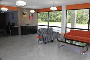 En modern entréhall med en receptionsdisk, en svartvit mönstrad soffa, en orange soffa och stora fönster med utsikt över grönområden.