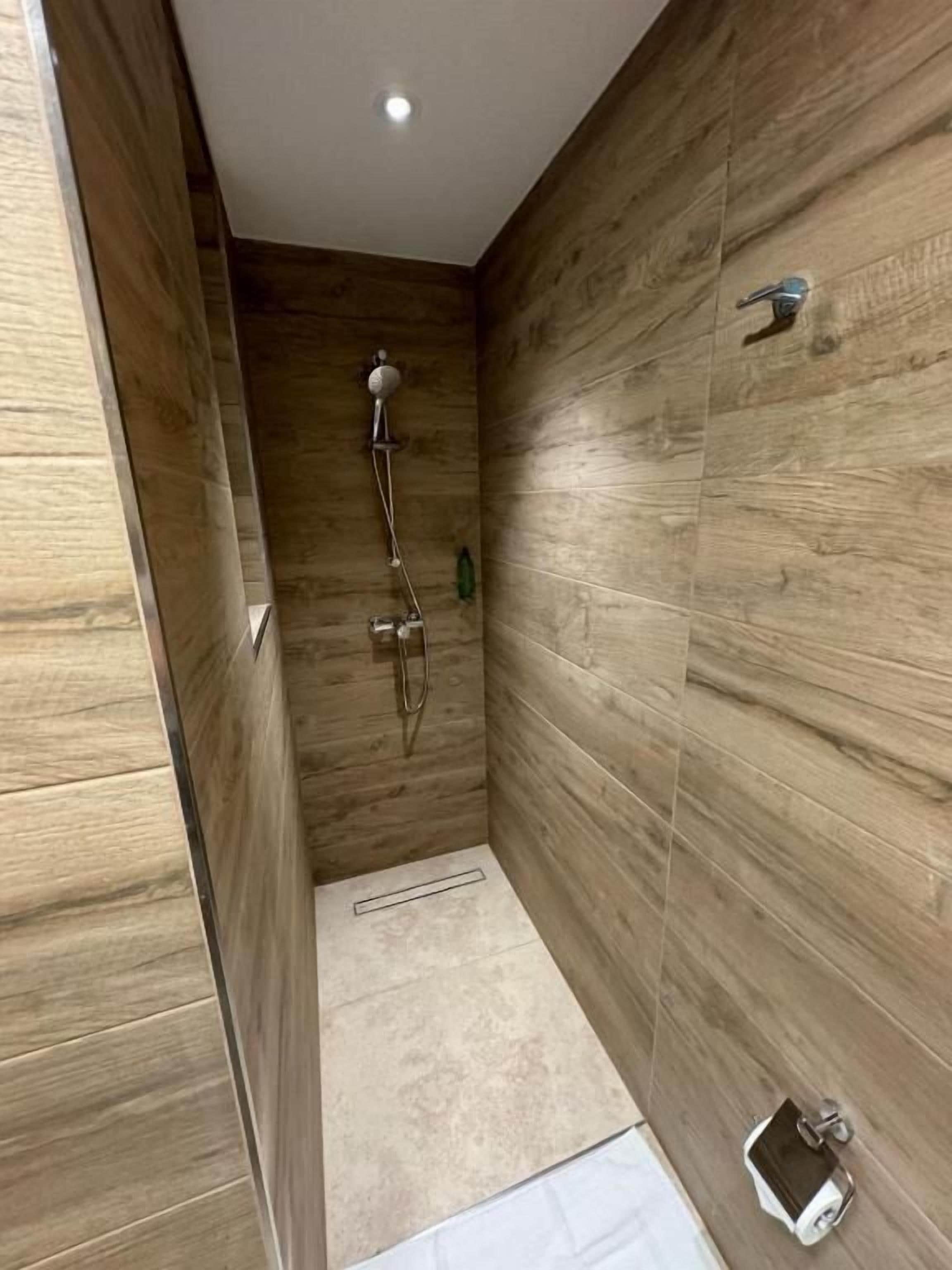 Un espace douche étroit aux murs recouverts de panneaux en bois, doté d’un pommeau de douche et d’un flexible de douche.