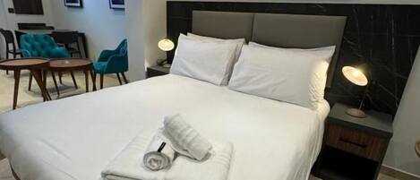 Una habitación de hotel con una cama grande, cabecera, mesita de noche con lámpara, un cuadro en la pared y una pequeña mesa con sillas.