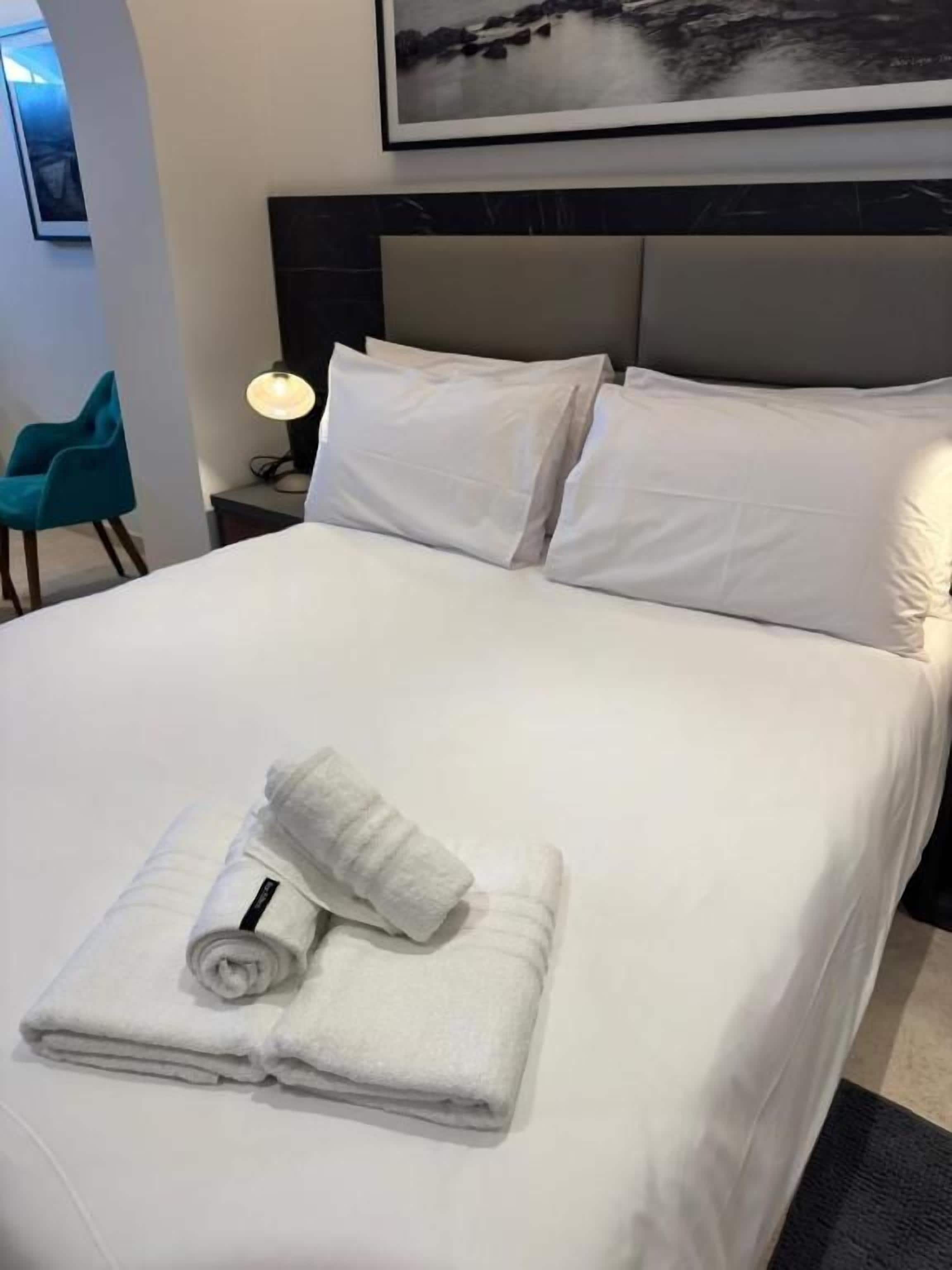 A neatly made bed with white linens and rolled towels, a headboard, and a framed picture above.