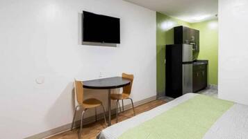 Una habitación de hotel con una cama, un escritorio con dos sillas, un televisor montado en la pared y una kitchenette con refrigerador.