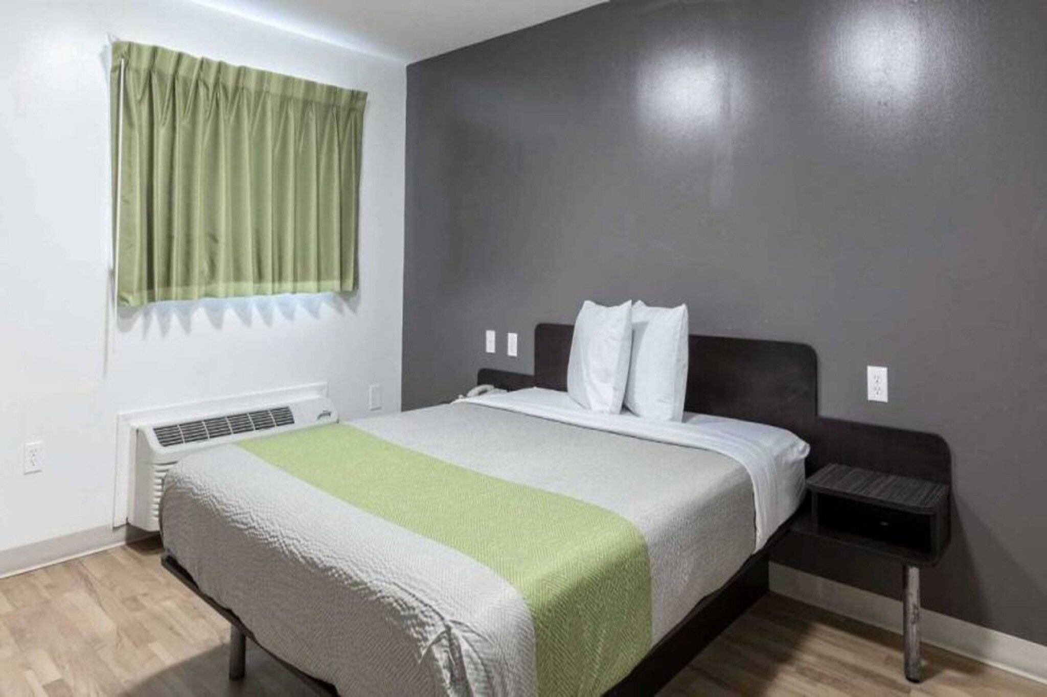 A bedroom with a bed, a bedside table, a wall-mounted heater, and a window with curtains.