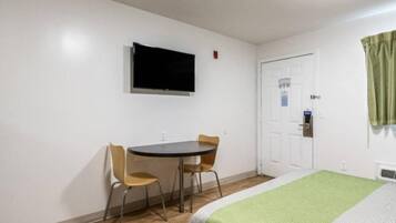 A small room with a bed, a desk with two chairs, a television mounted on the wall, and a door with a window.